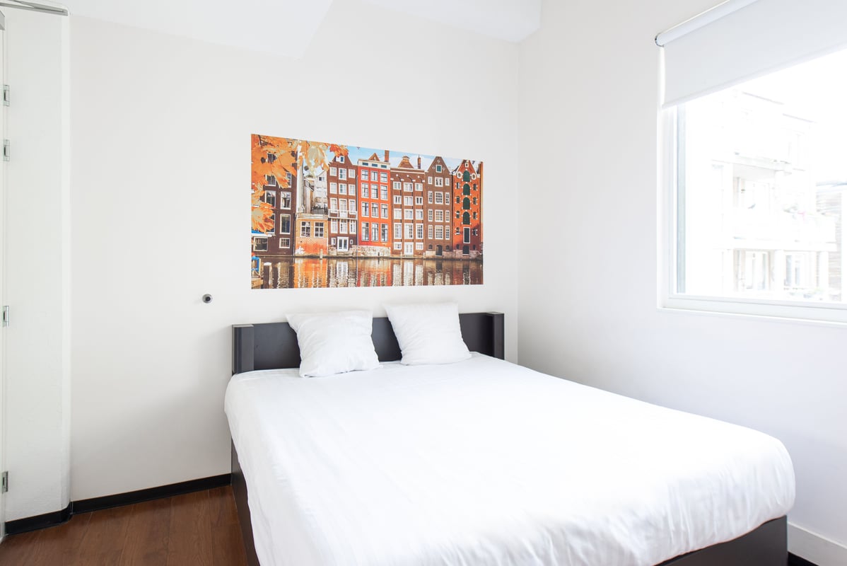 A clean, minimalist easyHotel room in Amsterdam City Centre South featuring a double bed with white linen and a large mural depicting colorful Amsterdam canal houses reflecting in a canal.