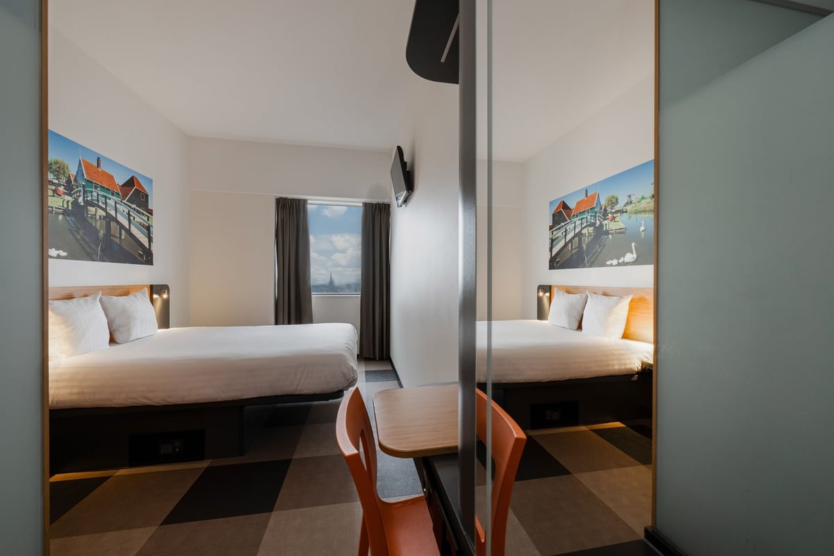Bright and modern easyHotel Amsterdam Zaandam twin room with two single beds, large wall art depicting a Dutch canal scene, and a city view from the window.