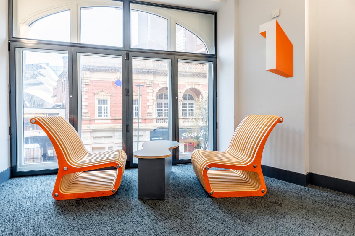 Modern lobby seating area at easyHotel Birmingham, featuring two orange and natural wood slatted lounge chairs facing a large window overlooking a city street. A small dark table is between the chairs, and an orange number '1' is on the wall.
