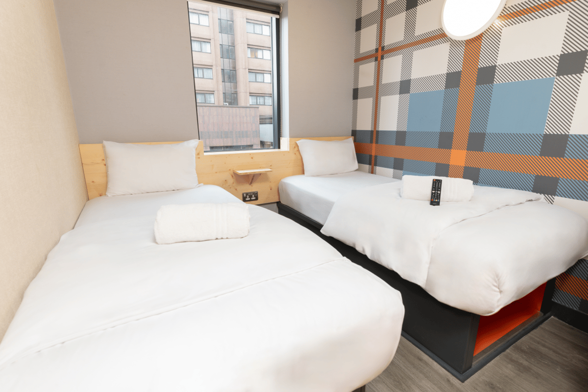 An easyHotel Glasgow twin room featuring two neatly made single beds with white bedding and towels. A prominent blue, orange, and grey plaid patterned accent wall is visible, alongside a window offering a city view.