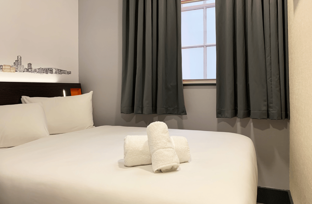 Clean, minimalist easyHotel room in Ipswich featuring a neatly made bed with white linen, two rolled white towels, a dark wooden headboard with integrated lighting, a cityscape wall decal, and a window with dark grey curtains.