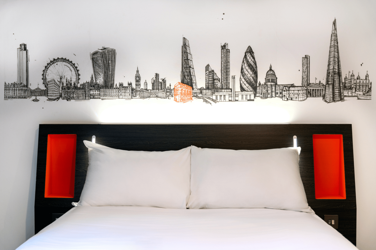 A bright easyHotel room featuring a comfortable bed with crisp white linens and a dark wood headboard. Above the bed, a detailed black and white mural depicts the iconic London skyline, with one building highlighted in orange.