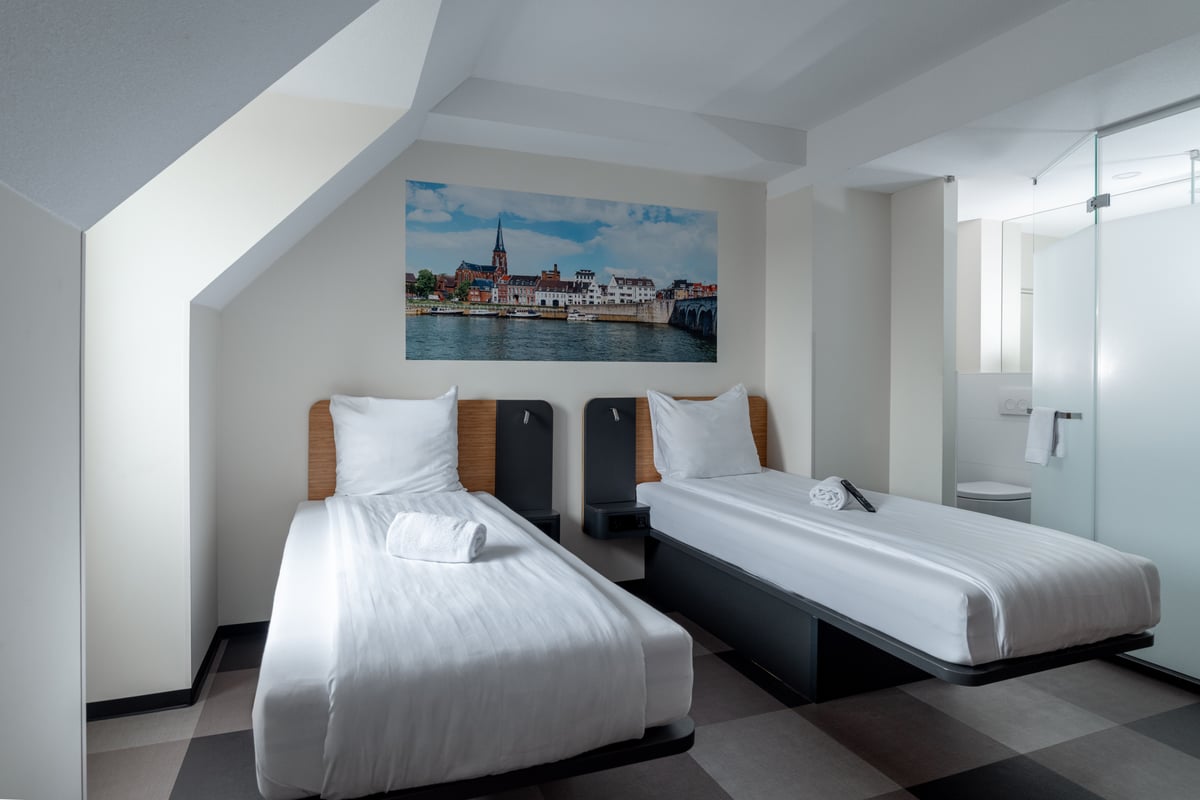A bright, modern easyHotel Maastricht room featuring two single beds with crisp white linens, a large wall mural depicting the Maastricht cityscape with a river and bridge, and a frosted glass bathroom enclosure.