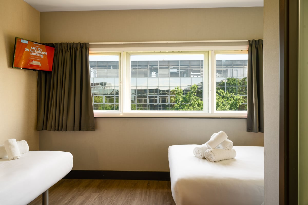 A bright and functional easyHotel twin room in Milton Keynes featuring two single beds with crisp white linen and folded towels, a wall-mounted TV displaying easyHotel branding, and a large window offering a view of a modern building and green trees.