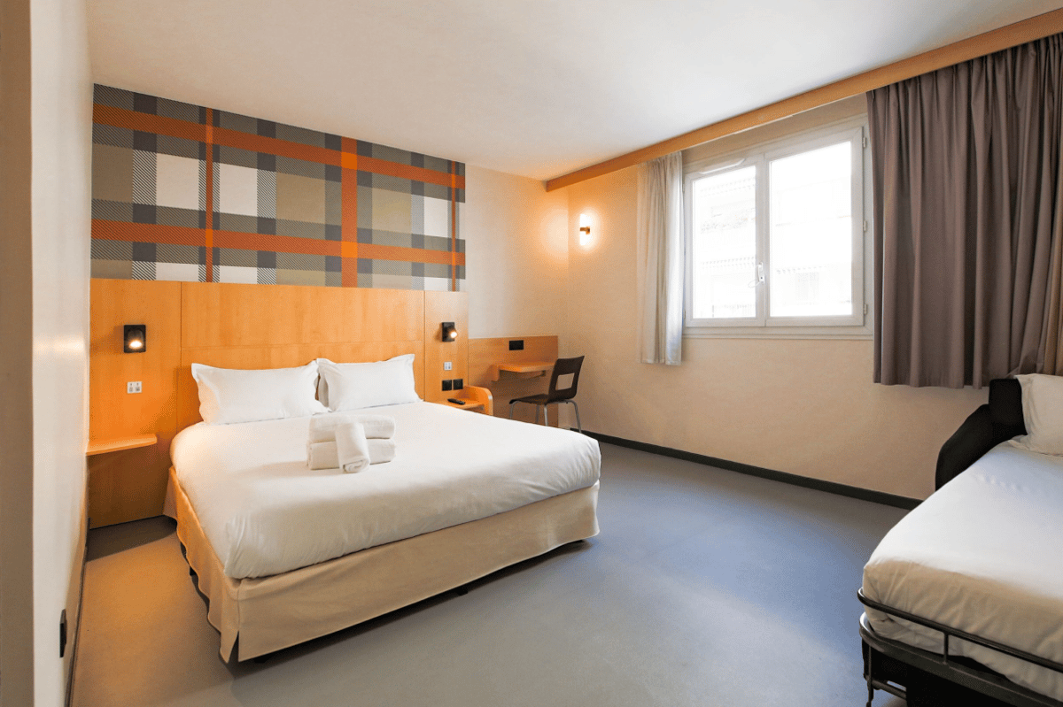 An easyHotel Nice Palais des Congres room featuring a double bed with white linen and towels, a light wood headboard, a vibrant orange and gray plaid accent wall, a functional desk with a chair, and a bright window.