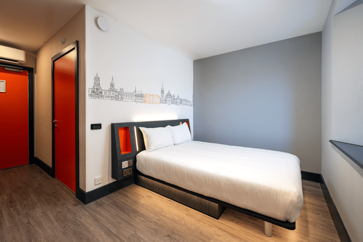ALT: easyHotel Oxford double room with a modern white bed, integrated headboard lighting, and a unique Oxford skyline mural in black and orange.