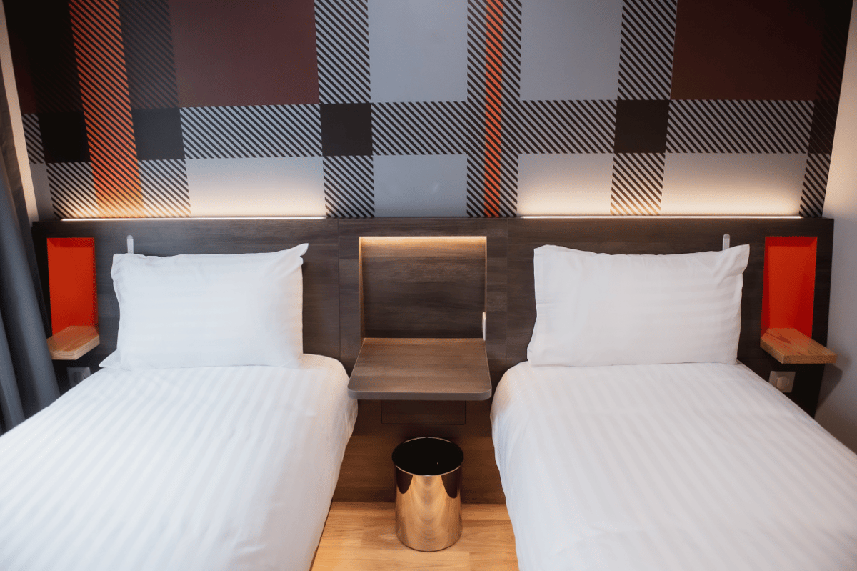 easyHotel Paris Nord Aubervilliers twin room featuring two single beds with white bedding, a shared central bedside table with integrated lighting, and a modern geometric wall pattern in black, grey, and orange above the beds.