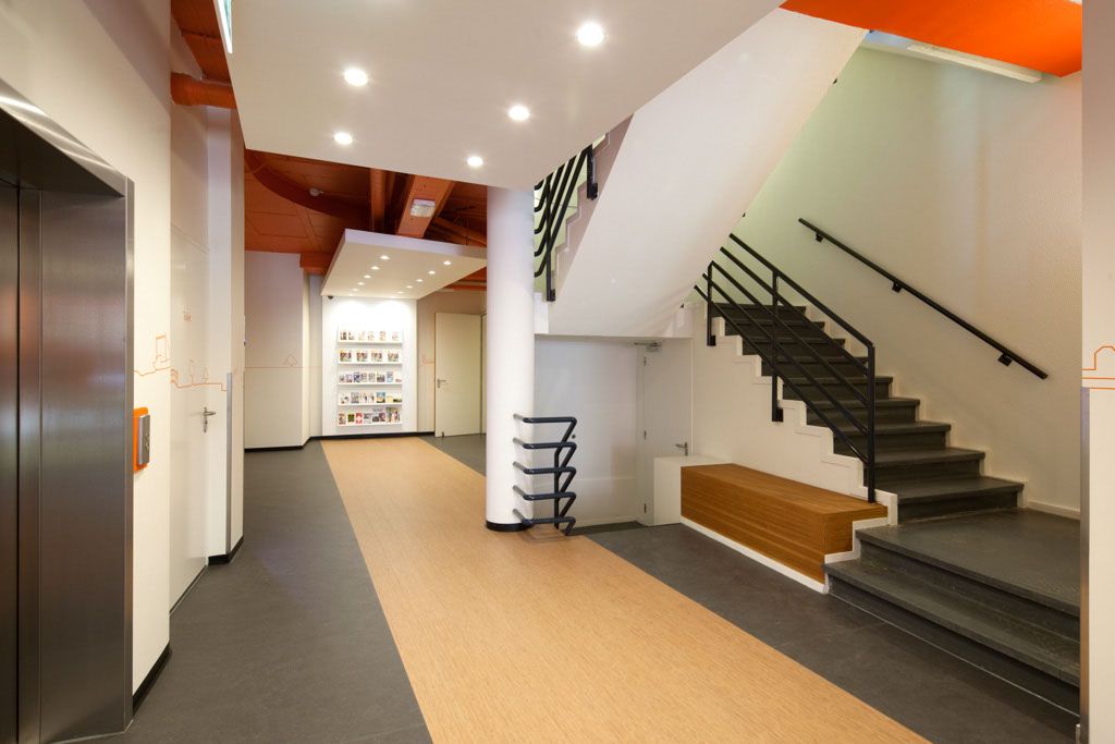 Modern easyHotel The Hague lobby featuring a prominent black-railed staircase, metallic elevator, and bright orange ceiling accents. A light wood-look floor strip leads past a central pillar towards a lit display area.