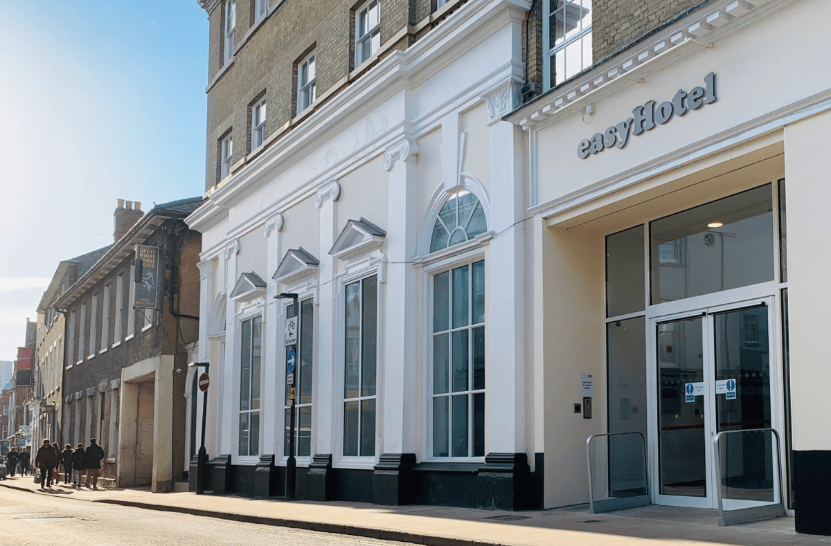 Exterior street view of easyHotel Ipswich on a sunny day. The cream-colored building features classical architectural details, large windows, and a prominent glass entrance. Pedestrians walk along the street outside the modern hotel.
