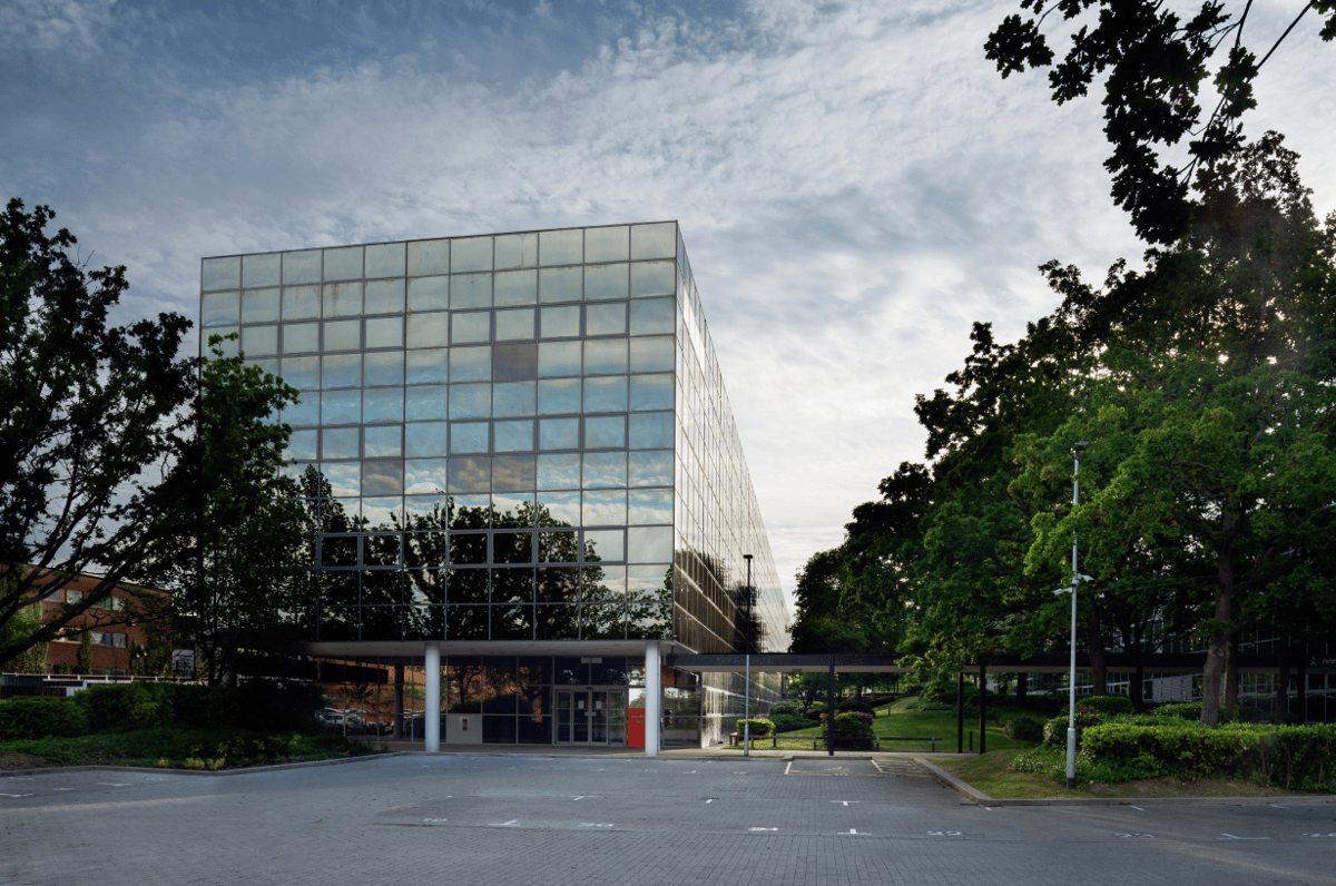 Exterior view of easyHotel Milton Keynes, a modern building with a reflective glass facade, a large parking lot, and surrounding green trees under a partly cloudy sky.