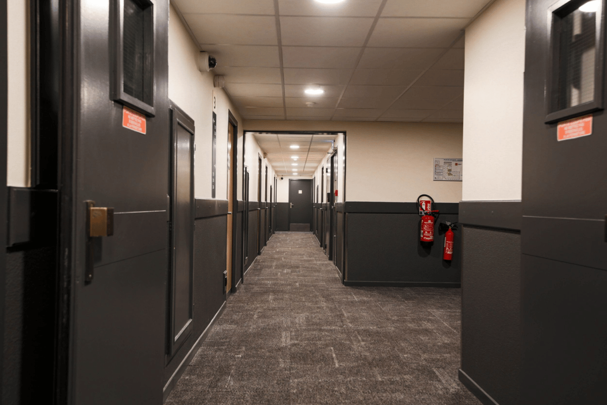 A long, well-lit hotel corridor at easyHotel Nice Palais des Congres, featuring dark gray doors, light beige upper walls, and a patterned dark gray carpet leading into the distance.