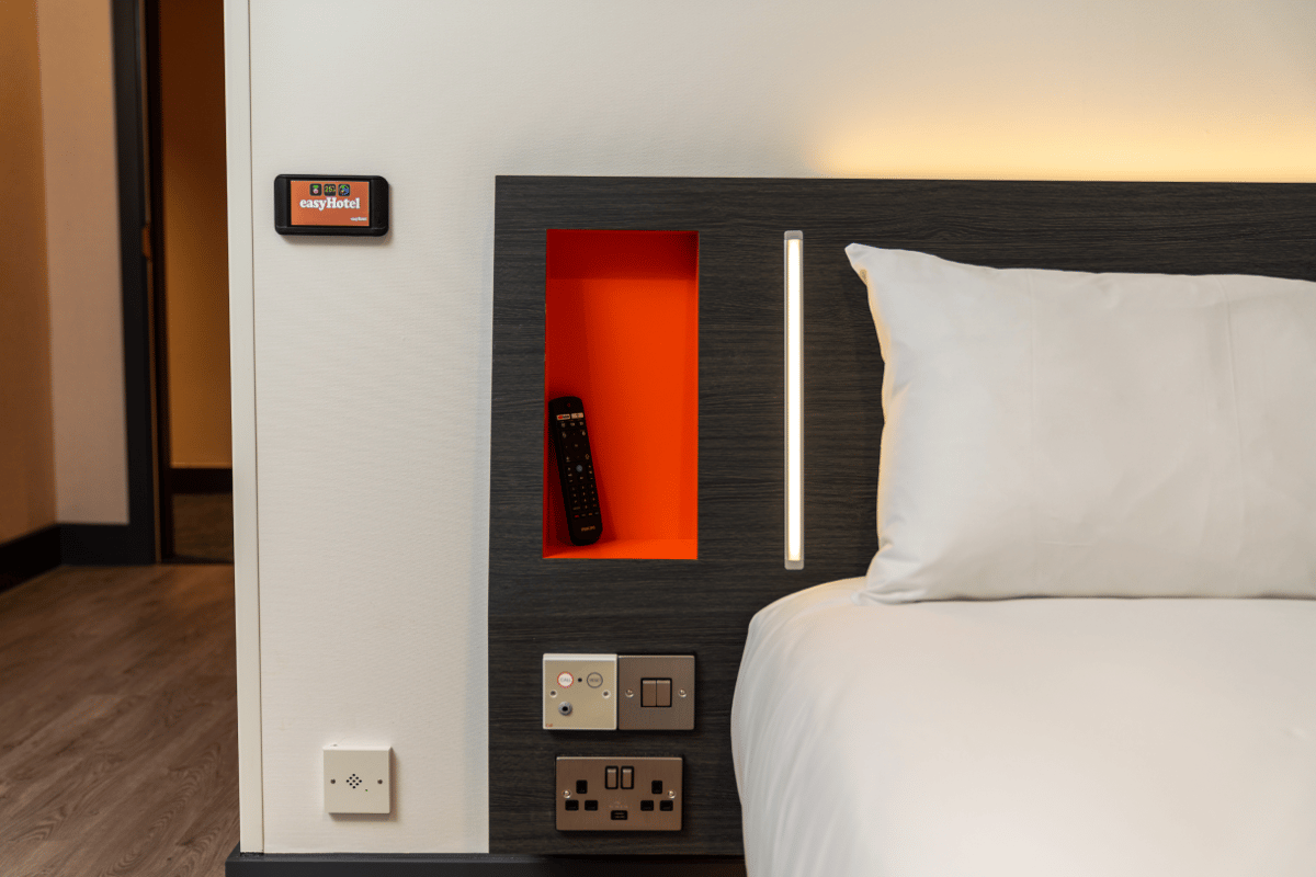 A modern and compact easyHotel Oxford room, featuring a white bed with a dark grey headboard, an orange recessed shelf holding a TV remote, built-in LED lighting, and accessible power outlets and USB ports near the bed.