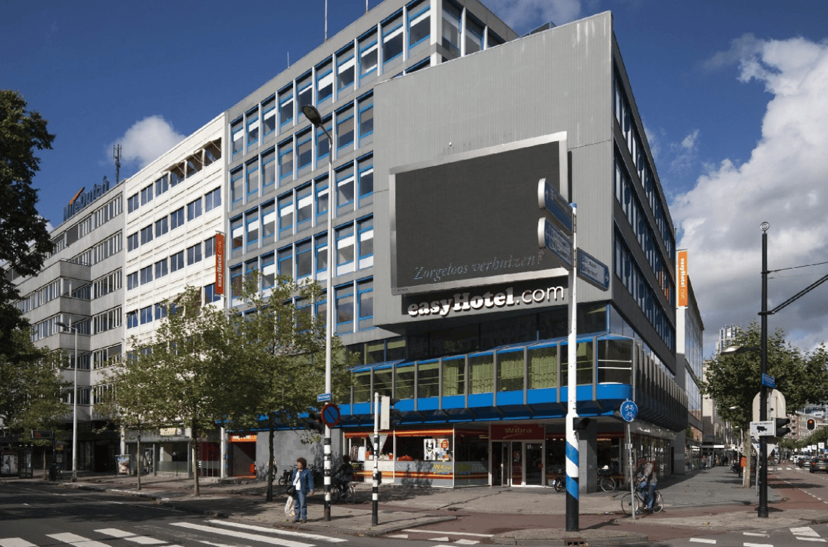 Exterior view of the easyHotel Rotterdam City Centre building, a modern multi-story hotel with a prominent easyHotel.com sign and a large digital display. The image captures the street-level facade under a blue sky, featuring a crosswalk, trees, and peopl
