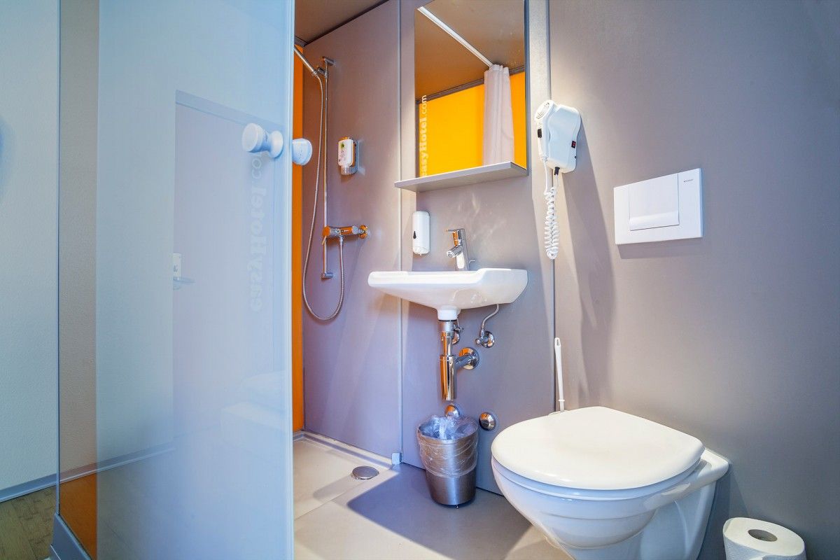 EasyHotel bathroom with integrated shower, compact sink, and toilet. The modern design features grey walls, bright orange accents, and essential fixtures like a mirror and hairdryer.