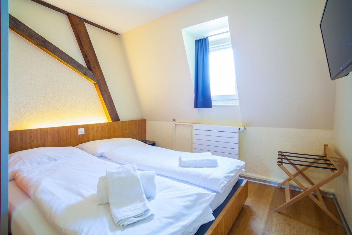 A bright, functional easyHotel room with two single beds, white linens, and towels. The room features a wooden headboard with accent lighting, exposed wooden ceiling beams, a dormer window with blue curtains, a wall-mounted TV, and a luggage rack on a woo