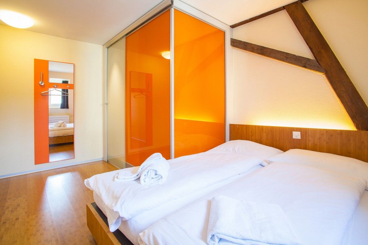 easyHotel room with a white bed, bright orange sliding doors, an orange wall mirror with hangers, a wooden headboard, and exposed wooden ceiling beams.