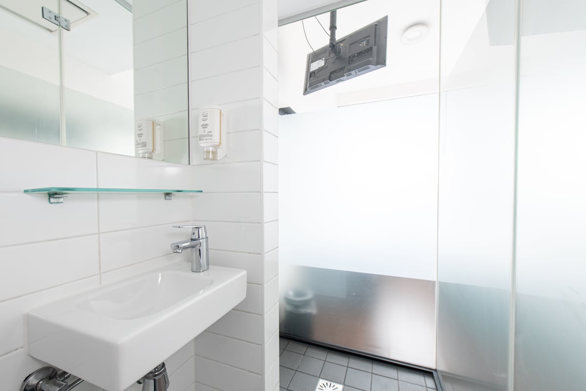 A modern, compact easyHotel Amsterdam City Centre South bathroom featuring a white rectangular sink, mirror, and a separate shower area with a unique ceiling-mounted flat-screen TV.