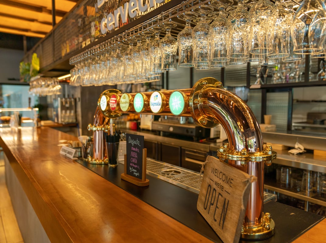A brightly lit bar counter at easyHotel Barcelona, featuring a prominent golden beer tap system, rows of clean glasses hanging above, and a wooden "Welcome - we're OPEN" sign.