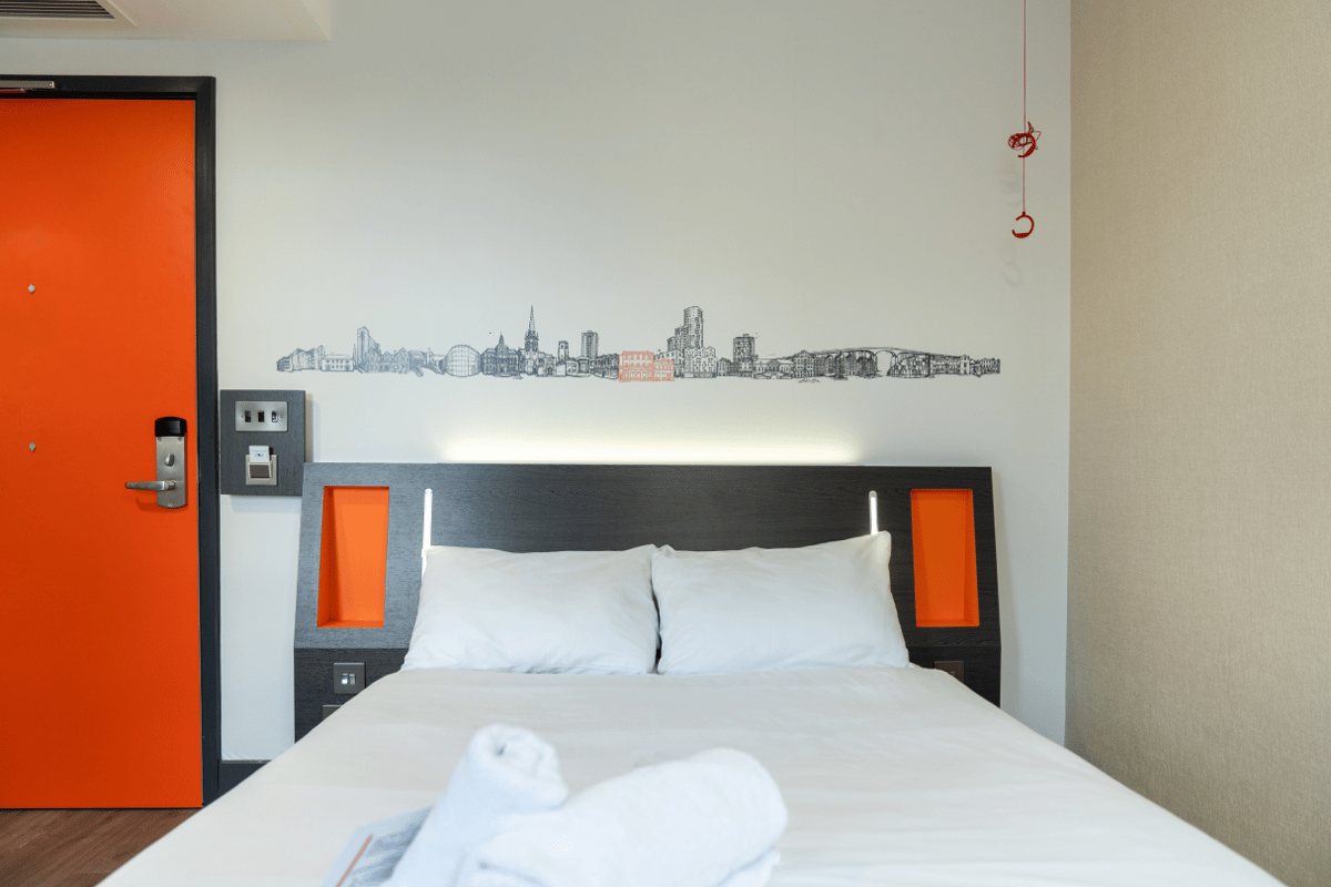 easyHotel Ipswich double bedroom featuring a white bed with a dark headboard and orange accents, integrated lighting, and an orange door. A black and white Ipswich city skyline mural with an orange highlight is on the wall above the bed.