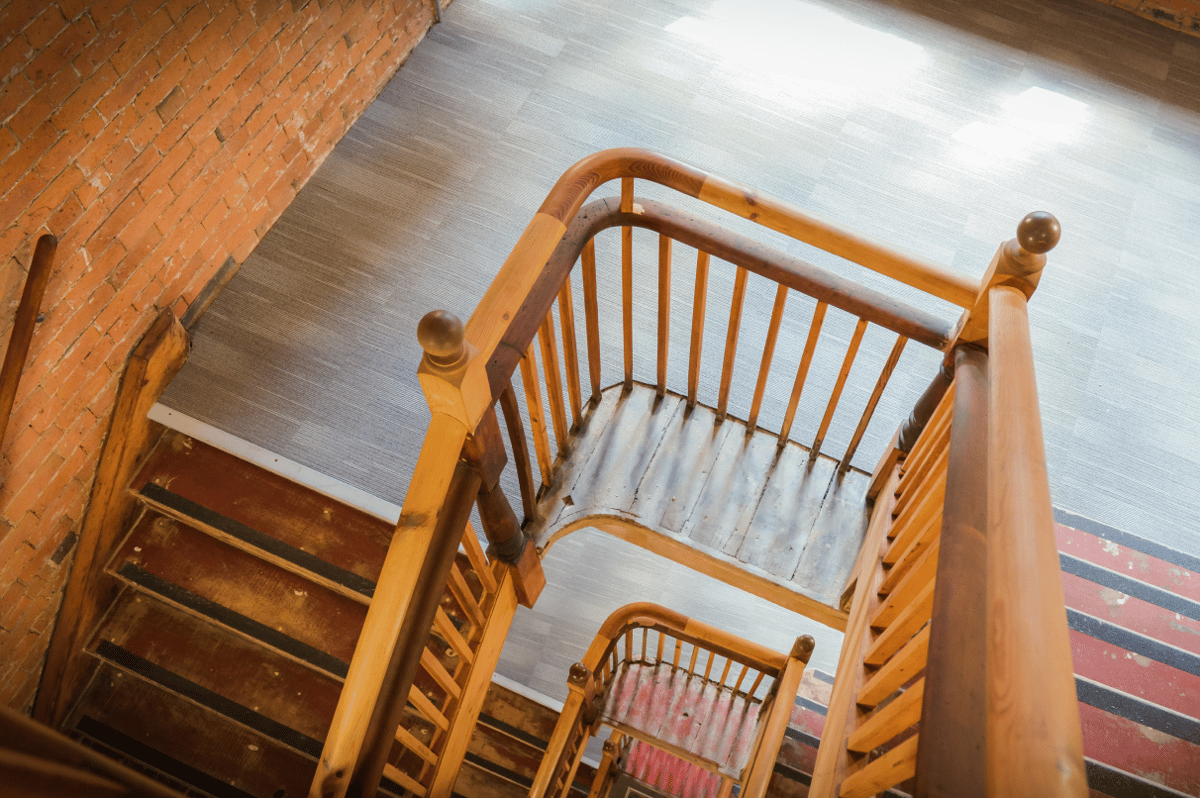 Overhead view of a winding wooden staircase with worn red steps, dark wood banisters, and an exposed brick wall leading to a modern gray carpeted landing at easyHotel Manchester.