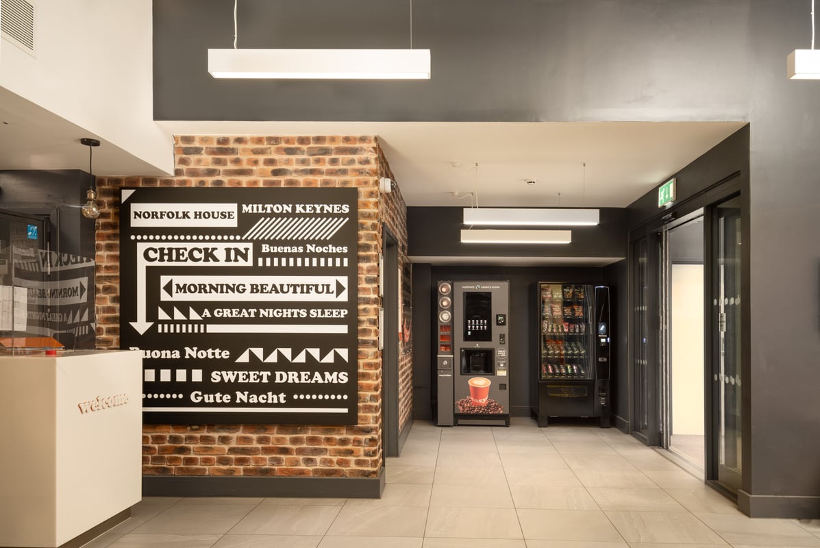 Modern easyHotel Milton Keynes lobby and reception area featuring a welcome desk, exposed brick wall with a large black and white check-in sign, and two self-service vending machines.