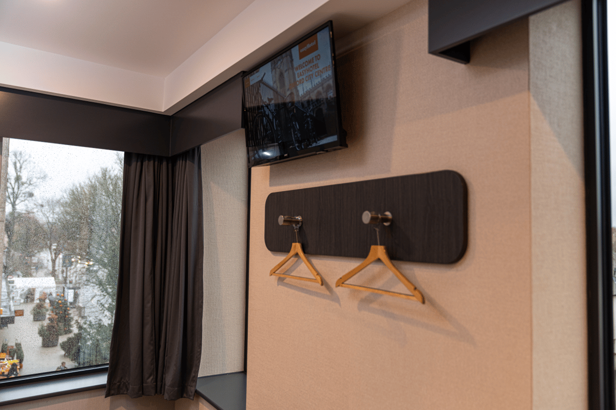 easyHotel Oxford room interior featuring a wall-mounted TV displaying a welcome message, a dark wood coat hanger rack with two wooden hangers, and a large window overlooking a city street with trees and an outdoor market setup.