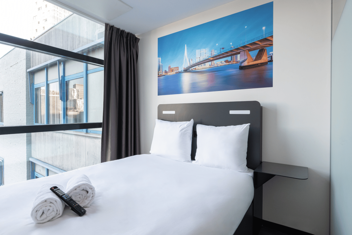 Modern easyHotel Rotterdam City Centre double room featuring crisp white bedding, a large window with city views, and a prominent wall print of the iconic Erasmus Bridge above the bed.