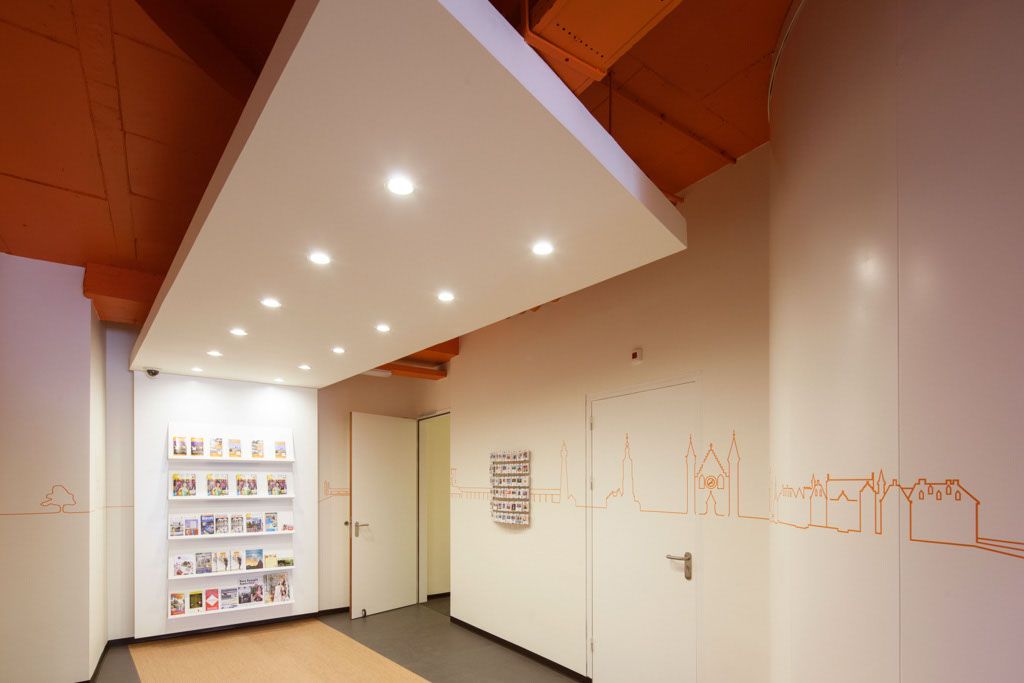 Interior of an easyHotel common area in The Hague, featuring white walls, orange ceiling details, and a suspended ceiling with bright spotlights. A wall-mounted display holds travel brochures, and an orange line drawing of The Hague city skyline decorates