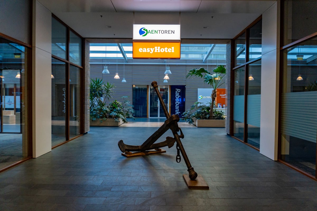 Lobby of easyHotel Zaandam, showcasing a large antique anchor sculpture in the center foreground, surrounded by modern glass walls, lush interior plants, and soft overhead lighting.
