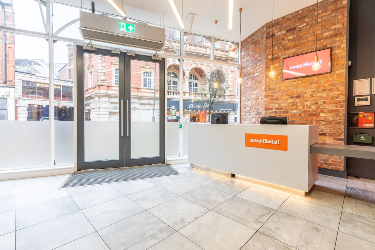 easyHotel Birmingham reception lobby with a white desk, orange easyHotel logo, exposed brick wall, and large windows looking out onto a bustling city street.