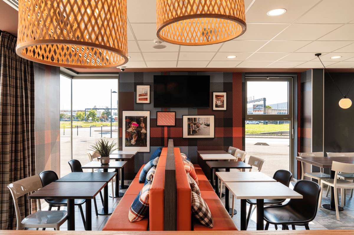 easyHotel CDG breakfast and lounge area with modern orange banquette seating, plaid pattern walls, and individual tables. Features bright natural light from large windows, wall-mounted art, and a casual, vibrant atmosphere.