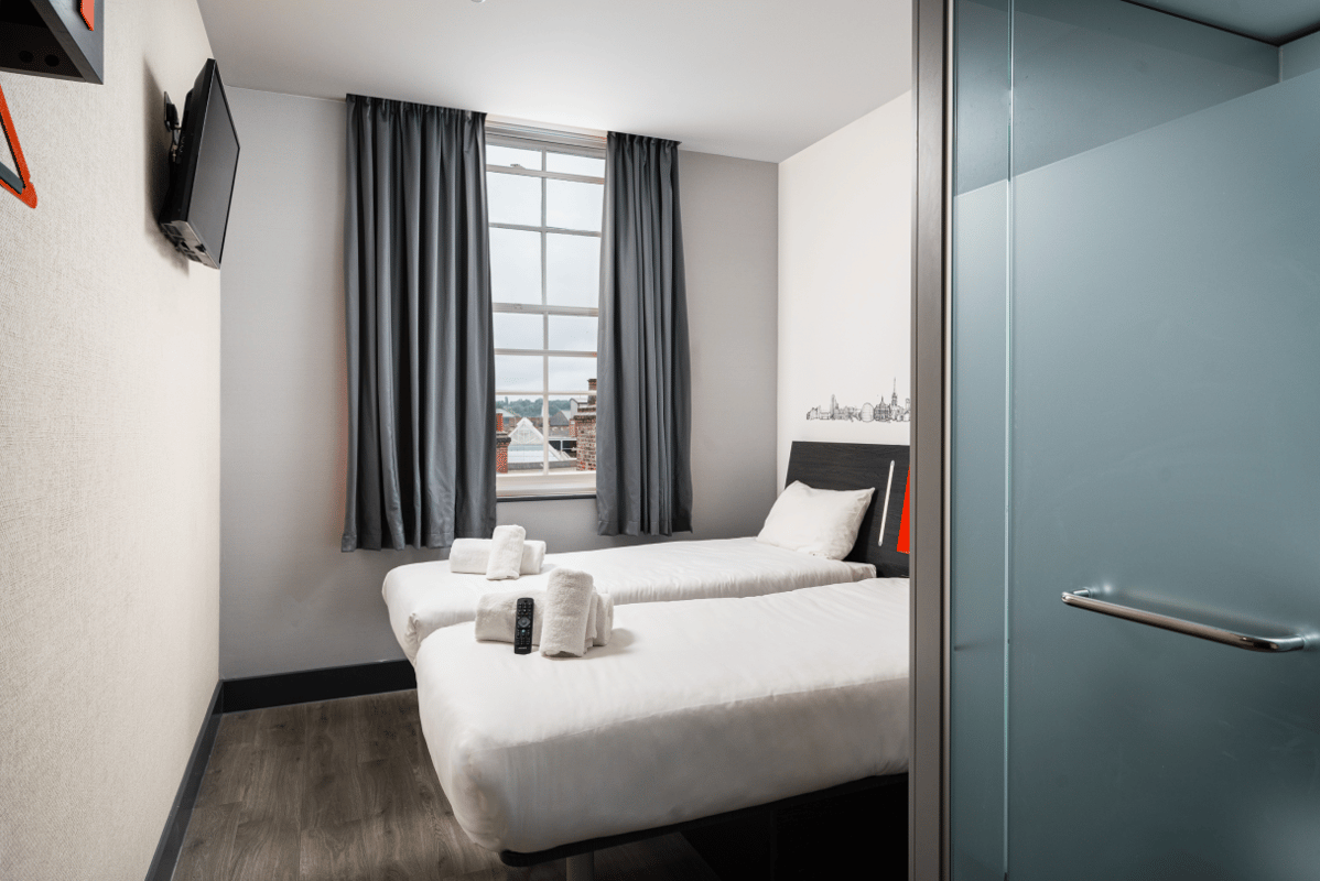 ALT: A bright and compact easyHotel twin room in Ipswich featuring two single beds with white linen, a wall-mounted TV, and a frosted glass bathroom door. White towels and a remote control are placed on one bed. The window offers a view of city rooftops.
