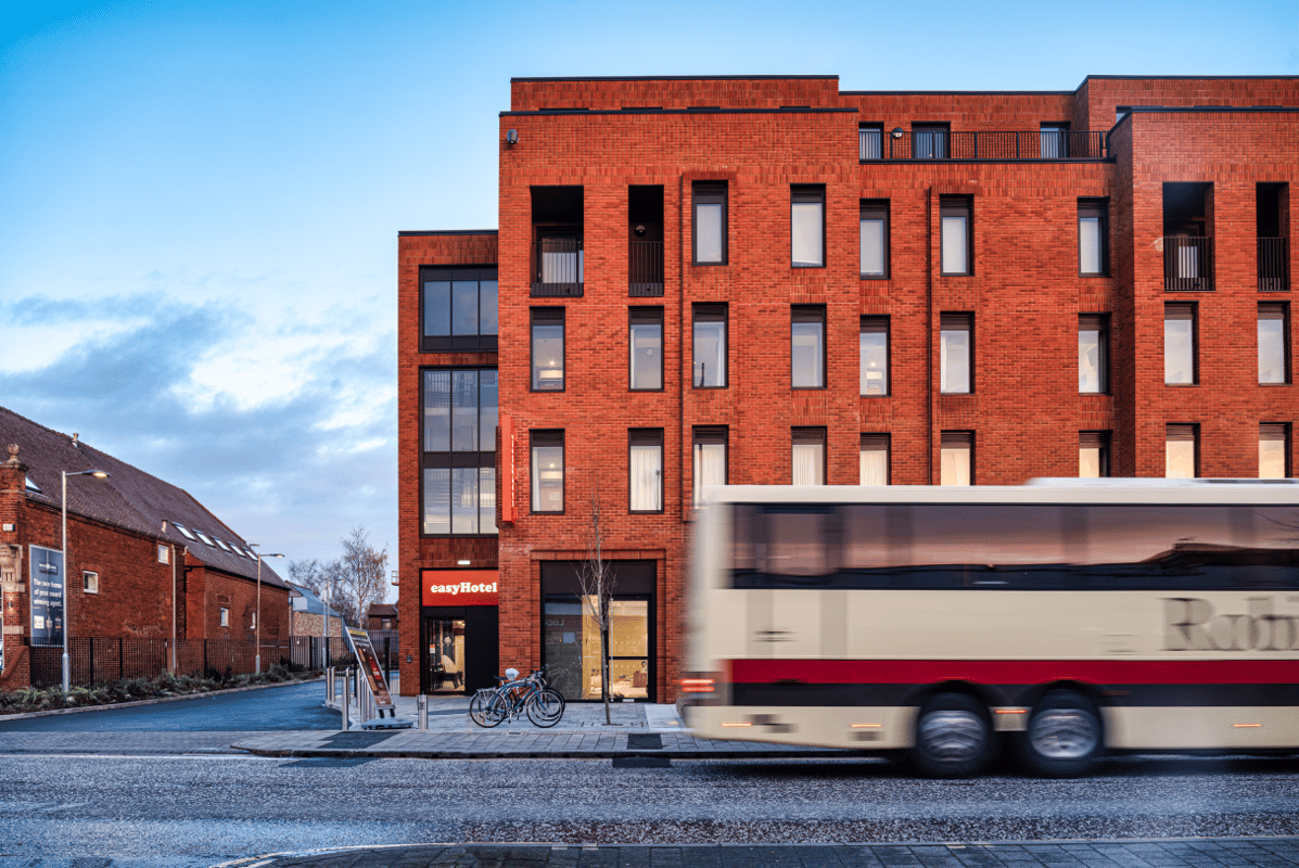 Exterior view of the modern red brick easyHotel building in Oxford at dusk, with a blurred bus passing by on the street and bicycles parked outside.
