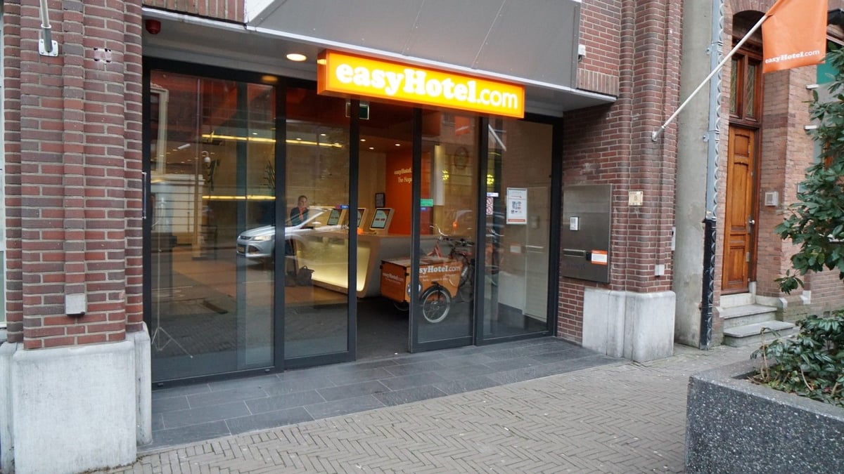 Exterior view of easyHotel The Hague, featuring its distinctive brick facade and glass entrance. A bright orange easyHotel.com sign is prominent above the doors, with an easyHotel branded cargo bike visible inside.