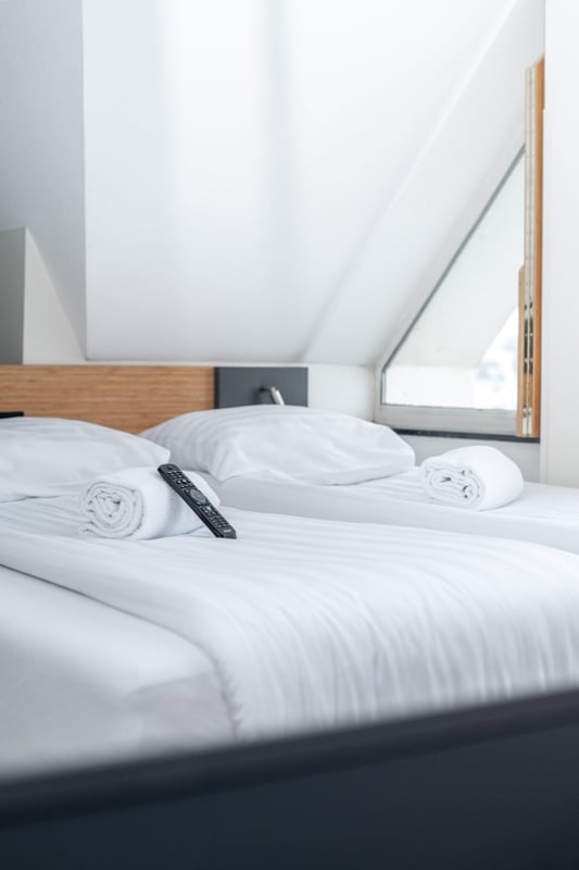 easyHotel Maastricht room with crisp white striped twin beds, a rolled towel, and a TV remote control. The clean, functional room features a sloped ceiling and an angled window.