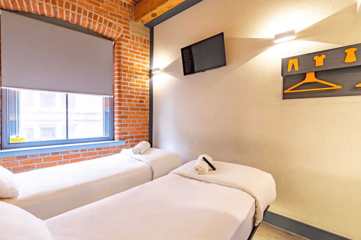 easyHotel Manchester room with two clean single beds, a wall-mounted television, an exposed brick wall with a large window, and a minimalist clothes rack with orange details.