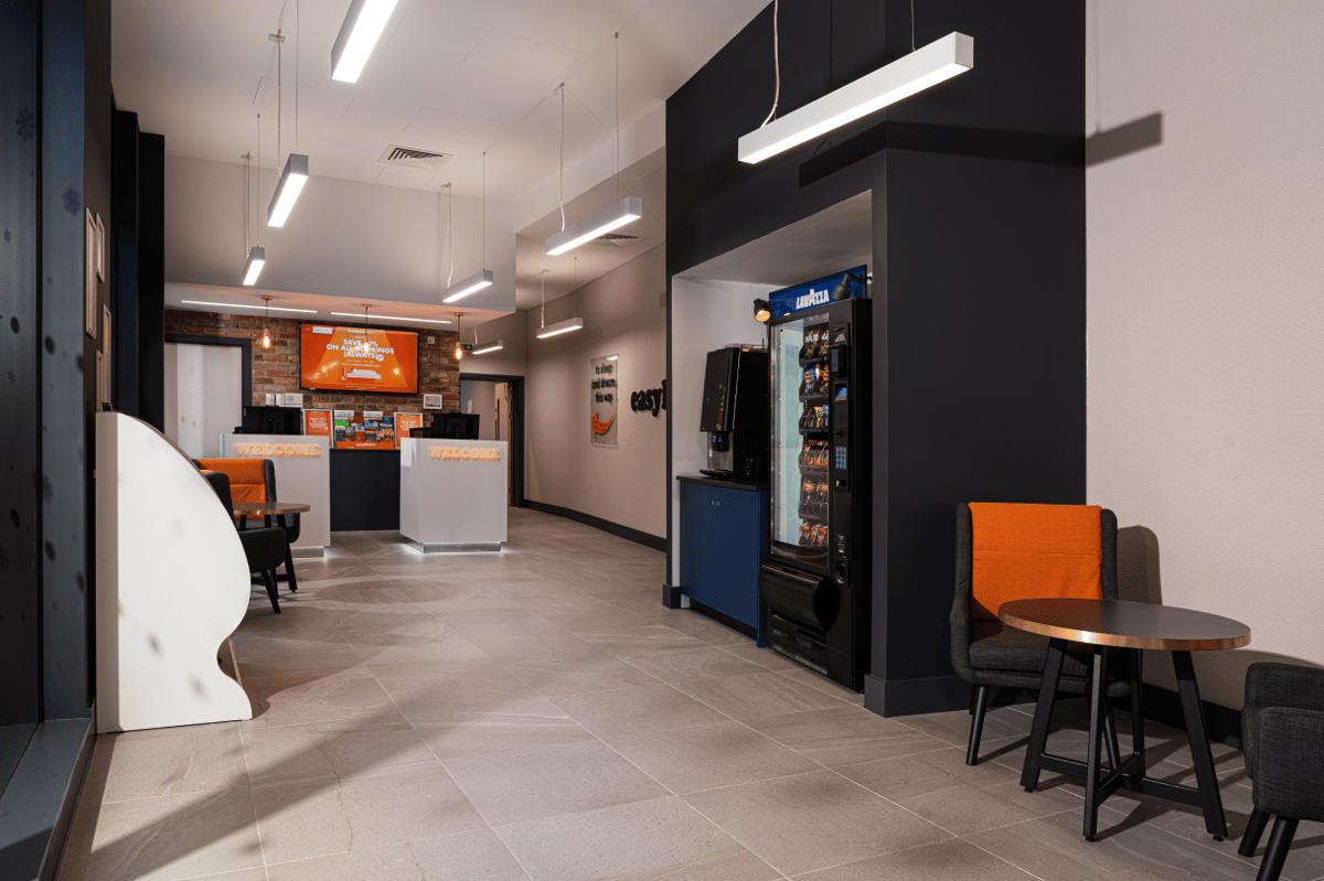 The welcoming, modern lobby of easyHotel Oxford, featuring a bright reception area with two 'WELCOME' desks, comfortable orange and grey seating, and a refreshment station with vending machines. The hotel's signature minimalist design is evident with ligh