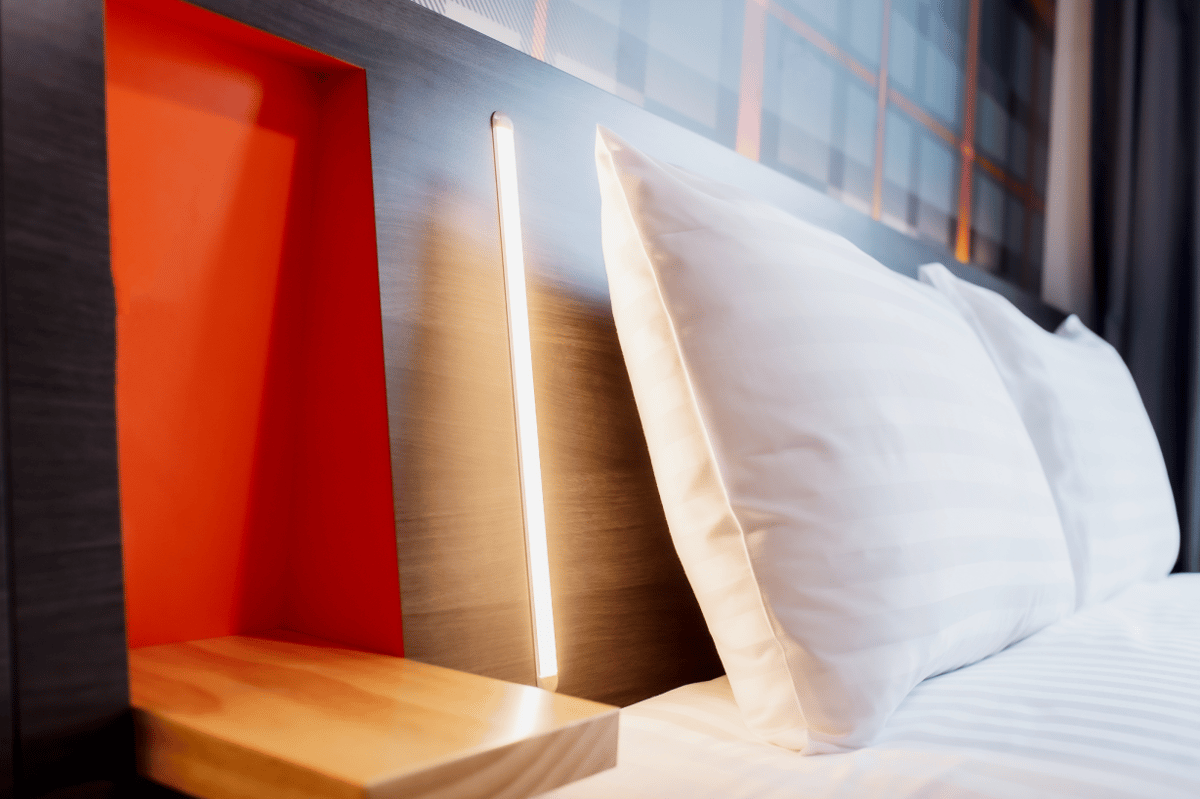 A close-up of a modern easyHotel room in Paris Nord Aubervilliers, highlighting the clean white bed with pillows, a dark headboard featuring an integrated vertical light strip, and a distinctive orange alcove with a wooden shelf.