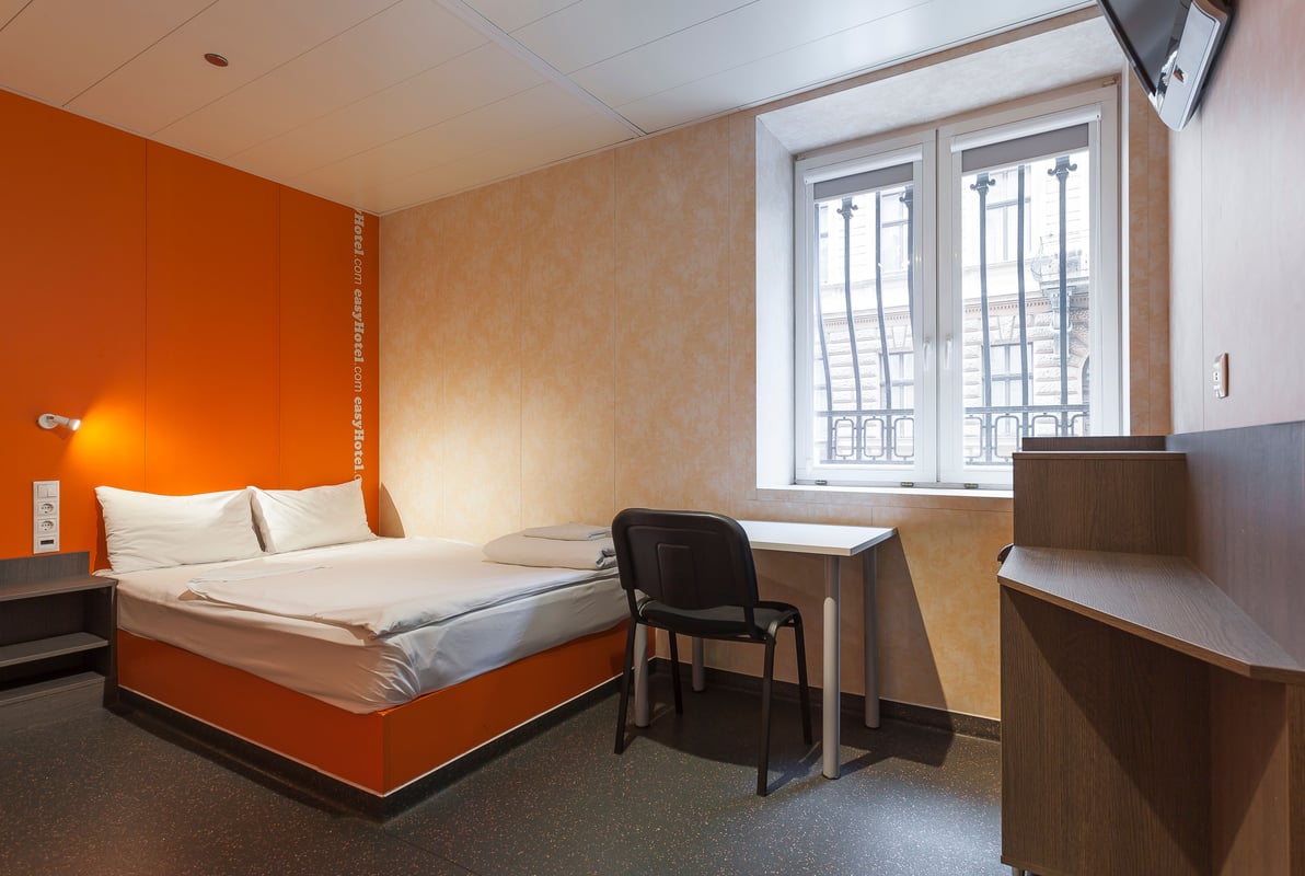 A compact easyHotel room featuring a double bed with crisp white linens against a vibrant orange accent wall. The room includes a minimalist desk with a black chair, a wall-mounted television, and a large window with ornate black iron bars, characteristic
