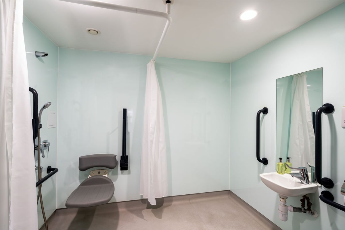 Accessible wet room in an easyHotel Liverpool City Centre, featuring light blue walls, grey floor, a fold-down shower seat, grab bars, and an accessible basin.