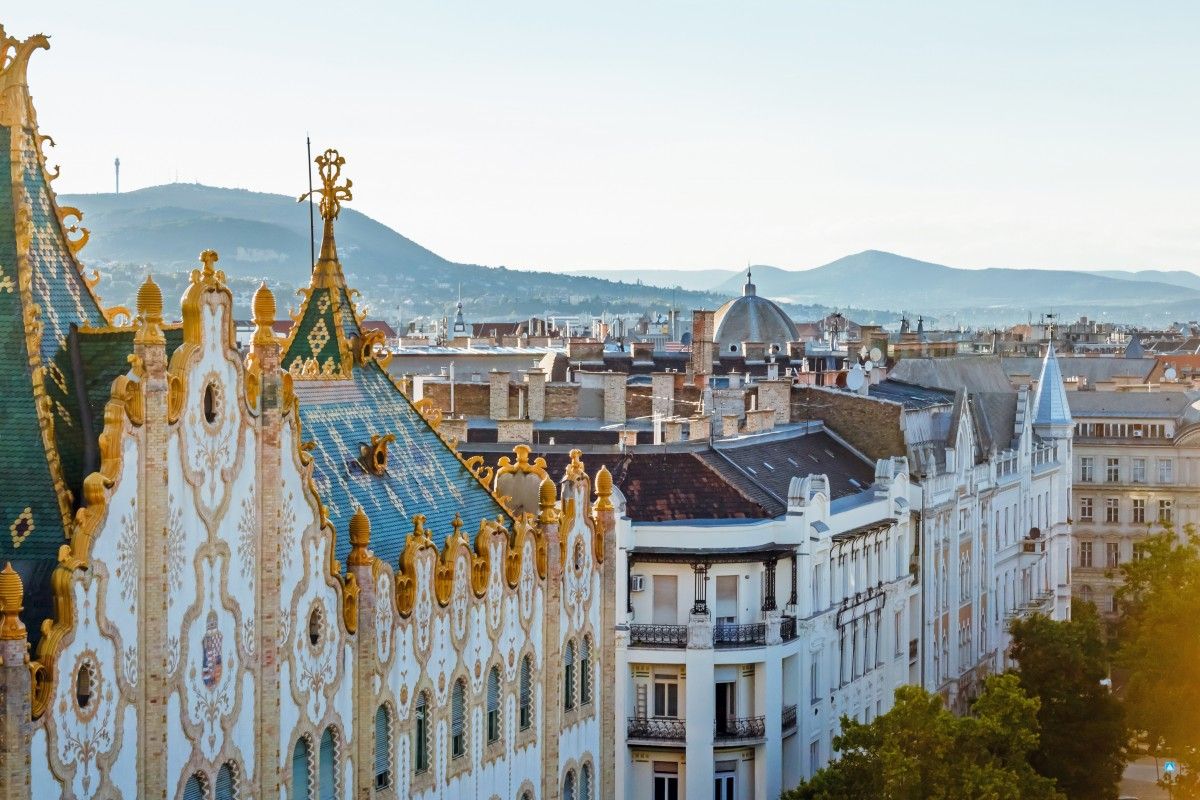 Budapest cityscape view featuring the ornate green and blue Zsolnay roof and decorative facade of the Museum of Applied Arts, with city buildings and distant hills under a clear sky.