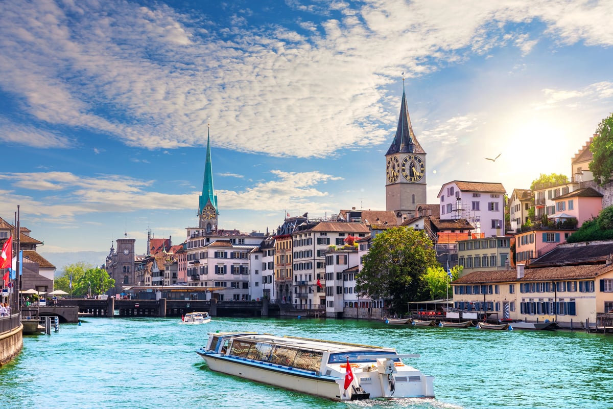 Zurich Old Town cityscape with a tour boat on the turquoise Limmat River, featuring historic riverside buildings, St. Peter's Church clock tower, and Fraumünster's green spire under a bright sky.