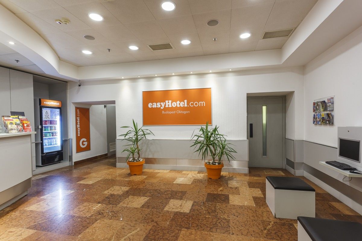 The modern and bright lobby of easyHotel Budapest Oktogon, featuring a reception desk, a refreshment vending machine, and a guest computer station, with the easyHotel.com sign prominently displayed on the wall.