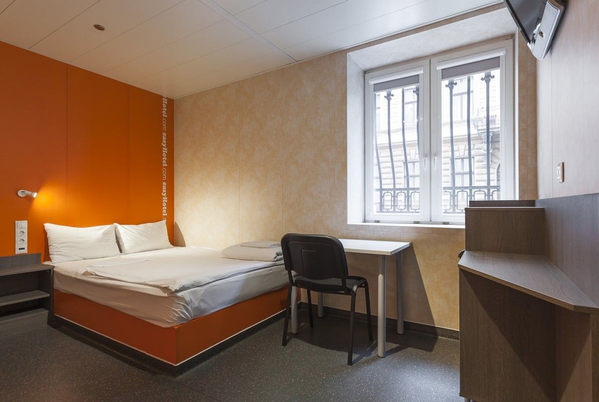 easyHotel compact room with an orange accent wall, a double bed on an orange base, a small desk, and a chair. A large window with ornate metal grilles offers an urban street view.