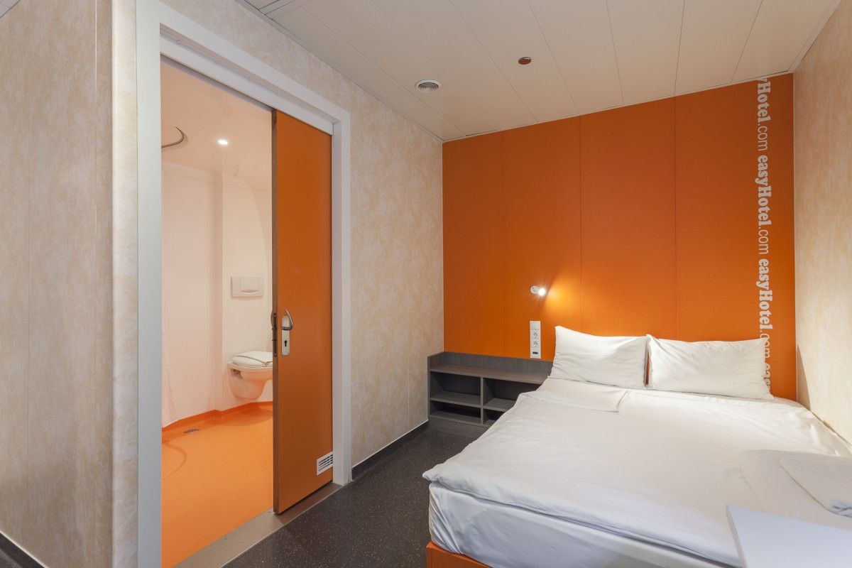 A compact easyHotel room with a double bed and white bedding against an orange accent wall. A partially open orange sliding door reveals the en-suite bathroom with an orange floor. The room features dark grey flooring and a simple built-in shelf.