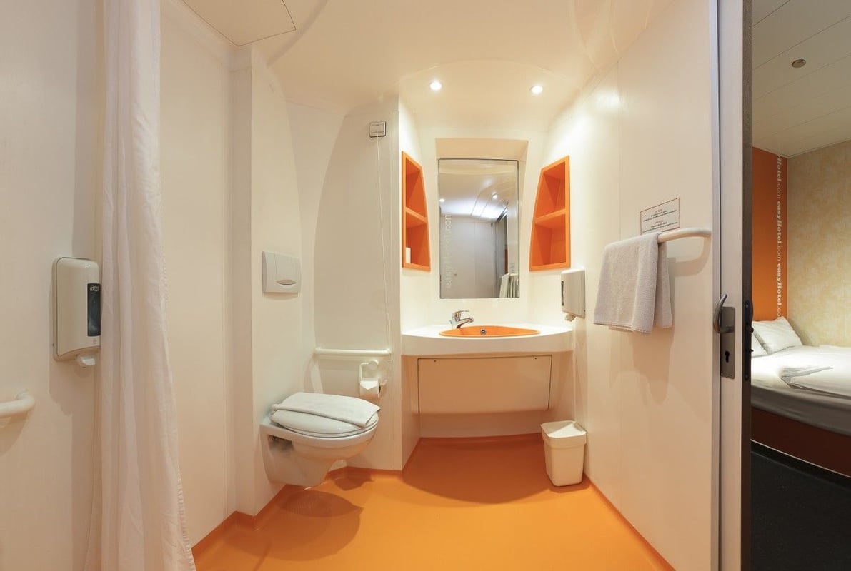 Modern easyHotel bathroom featuring a bright orange floor, orange sink, white toilet, and compact shower area, with a mirror and orange shelving. A sliver of the simple easyHotel bedroom is visible through the open doorway.