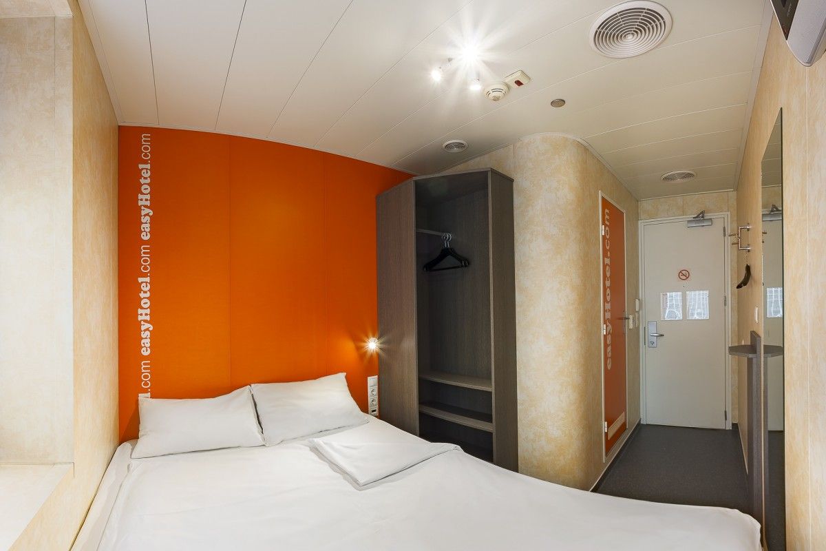 A compact easyHotel room with a white bed, bright orange accent wall featuring 'easyHotel.com' branding, and an open wardrobe. The room includes a modern white door, textured beige walls, and ceiling lights, ideal for budget hotel accommodation.