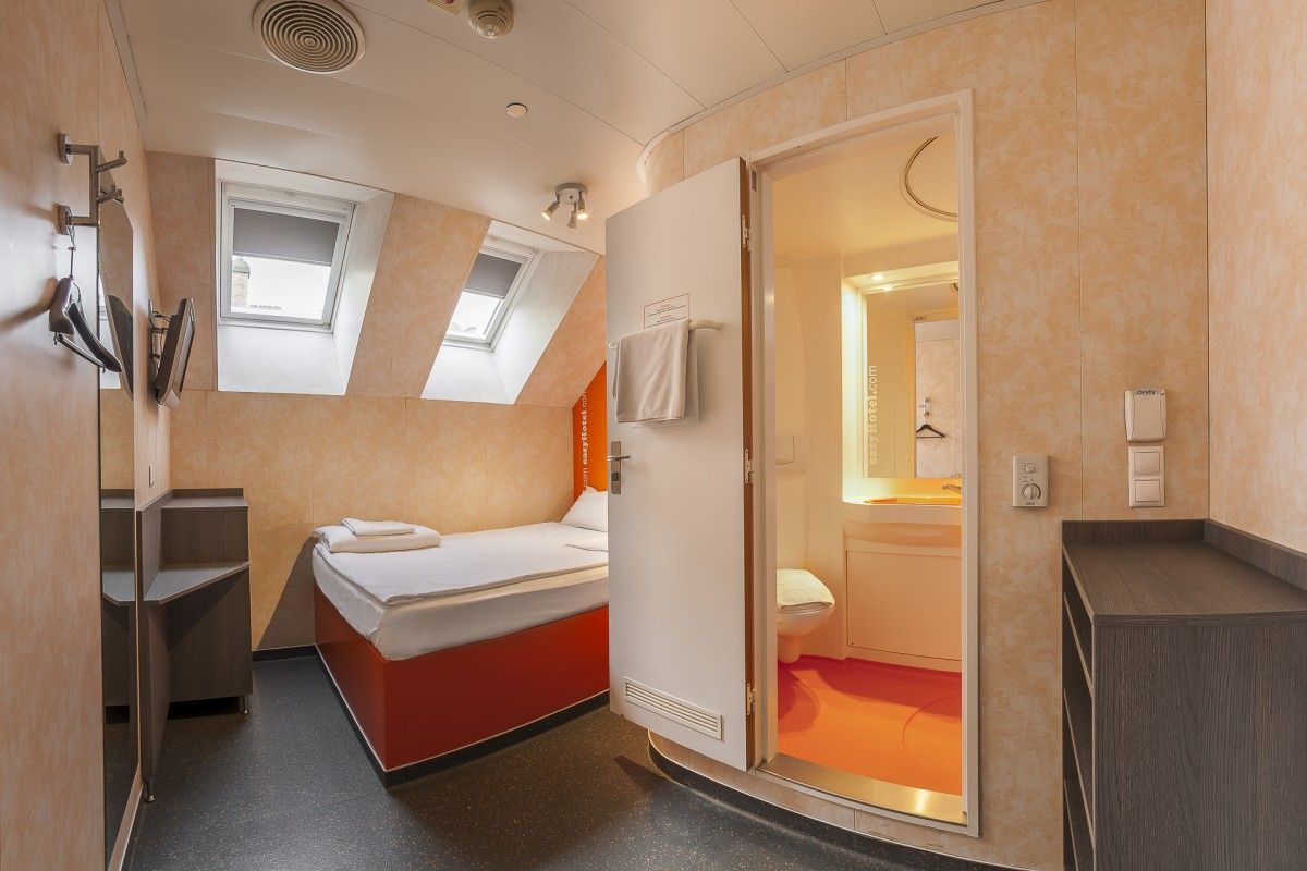 A compact and modern easyHotel room, featuring a single bed with a bright red base, light-toned walls, and a sloped ceiling with two skylight windows. An open door reveals a compact ensuite bathroom with a distinctive orange floor.