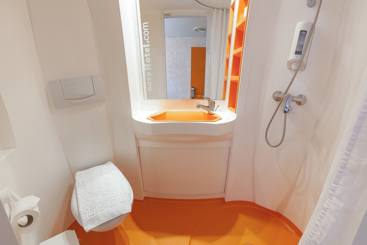 Compact easyHotel bathroom with bright orange non-slip floor, white walls, integrated toilet, small sink with mirror, and a shower area.