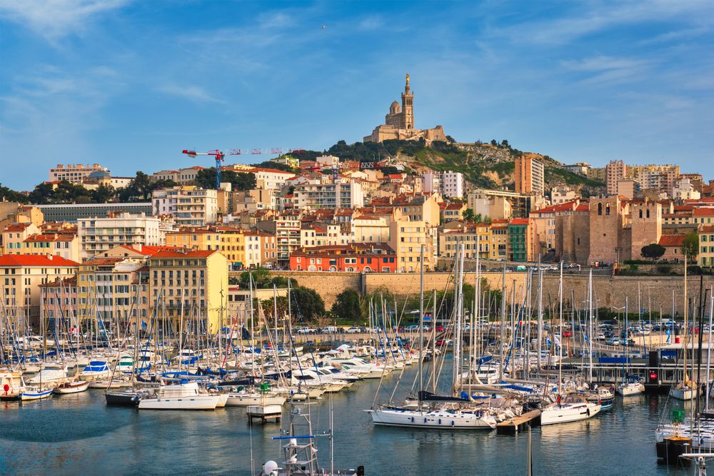 Vibrant Marseille Old Port cityscape with numerous sailboats and yachts, overlooked by the Notre-Dame de la Garde basilica on a hill under a clear blue sky.